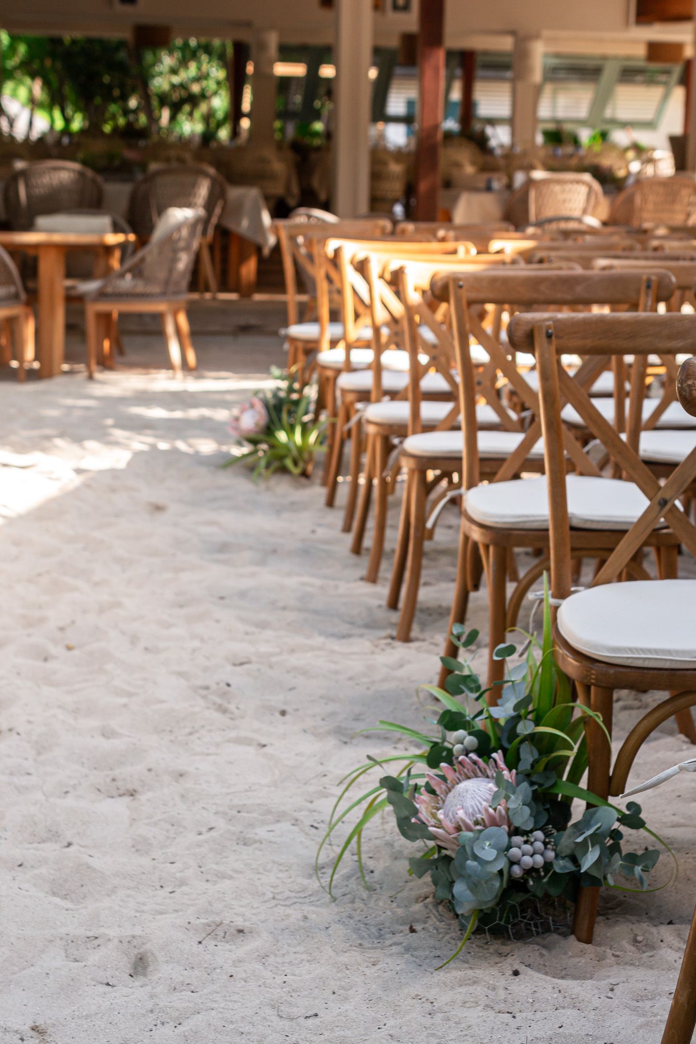 Rustic wooden chairs and tables arranged outdoors with small floral arrangements on a sandy ground.