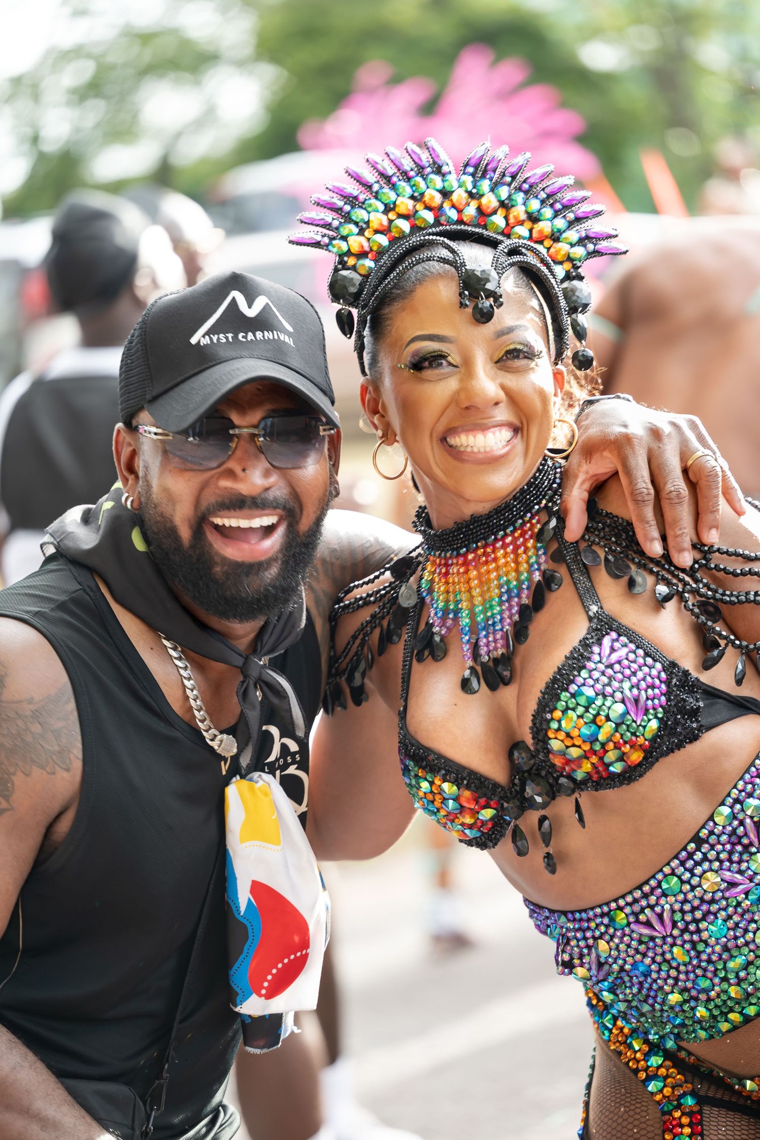Two people smiling at a Caribbean carnival, woman wearing colorful beaded costume and headdress, man in black cap.