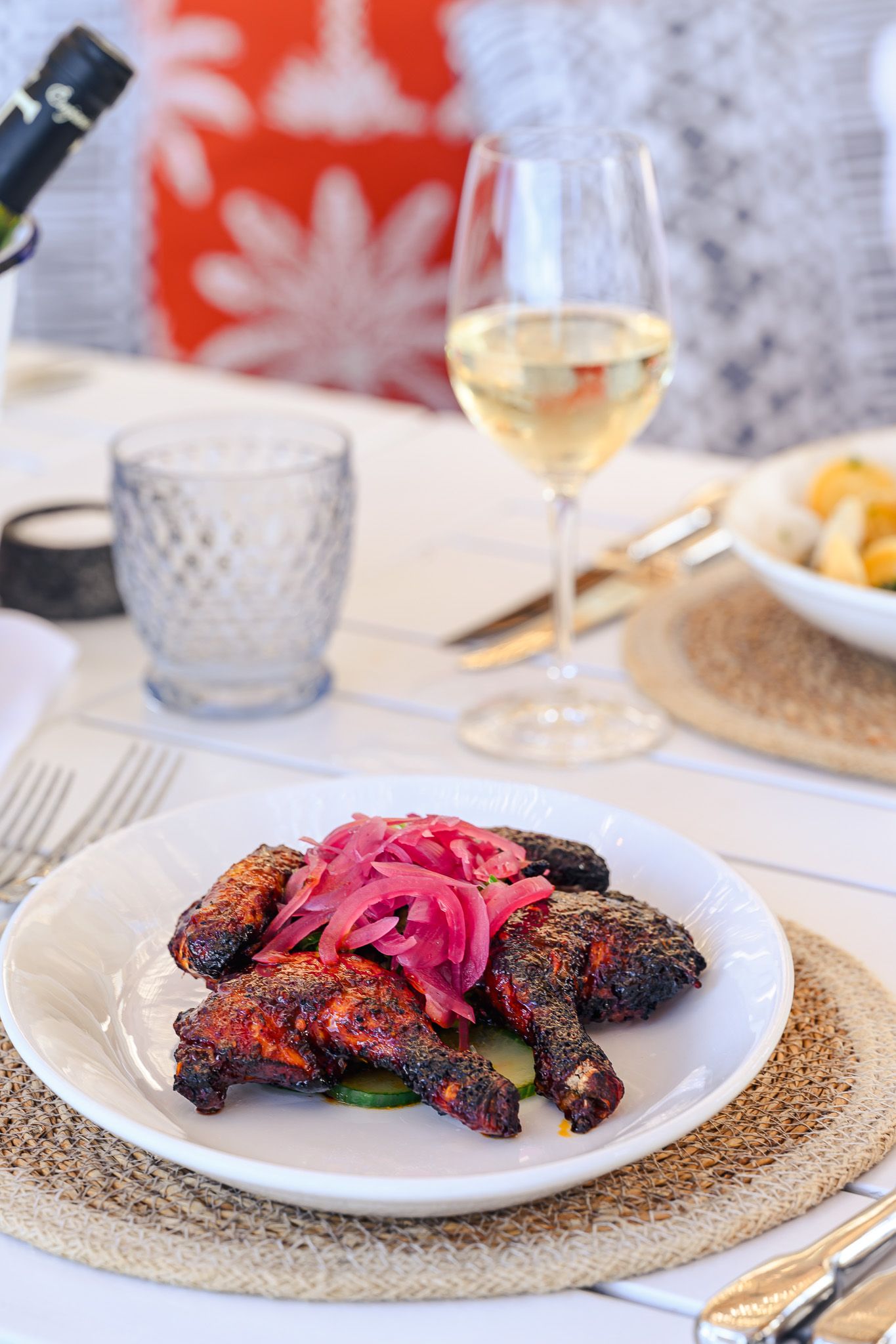 Grilled meat dish served on white plates with bright pink pickled garnish and woven placemats at an elegant table setting.