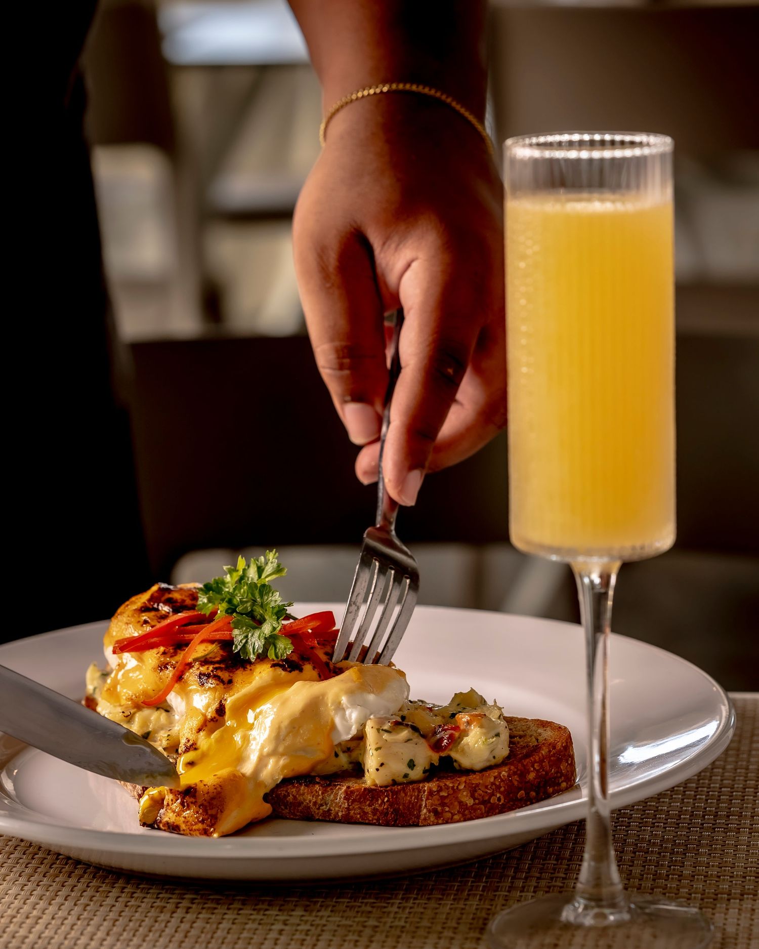 Hand forking into eggs benedict with hollandaise sauce on toast, served with a mimosa in a champagne flute.