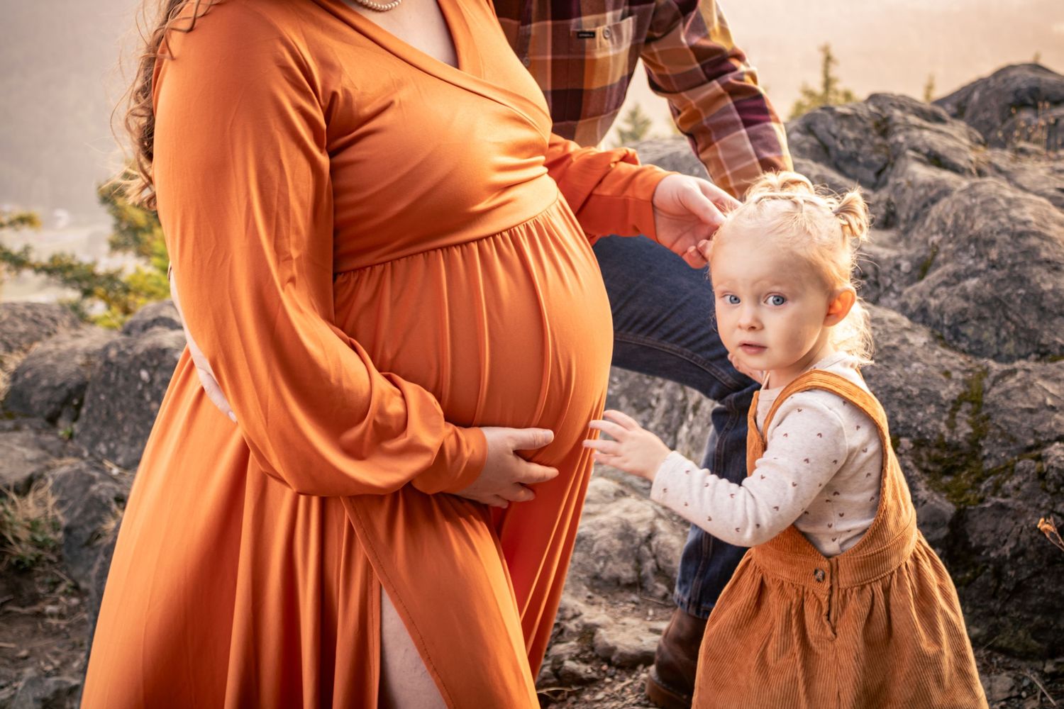 Pregnant woman in orange dress with toddler girl touching her baby bump during outdoor maternity photo session.