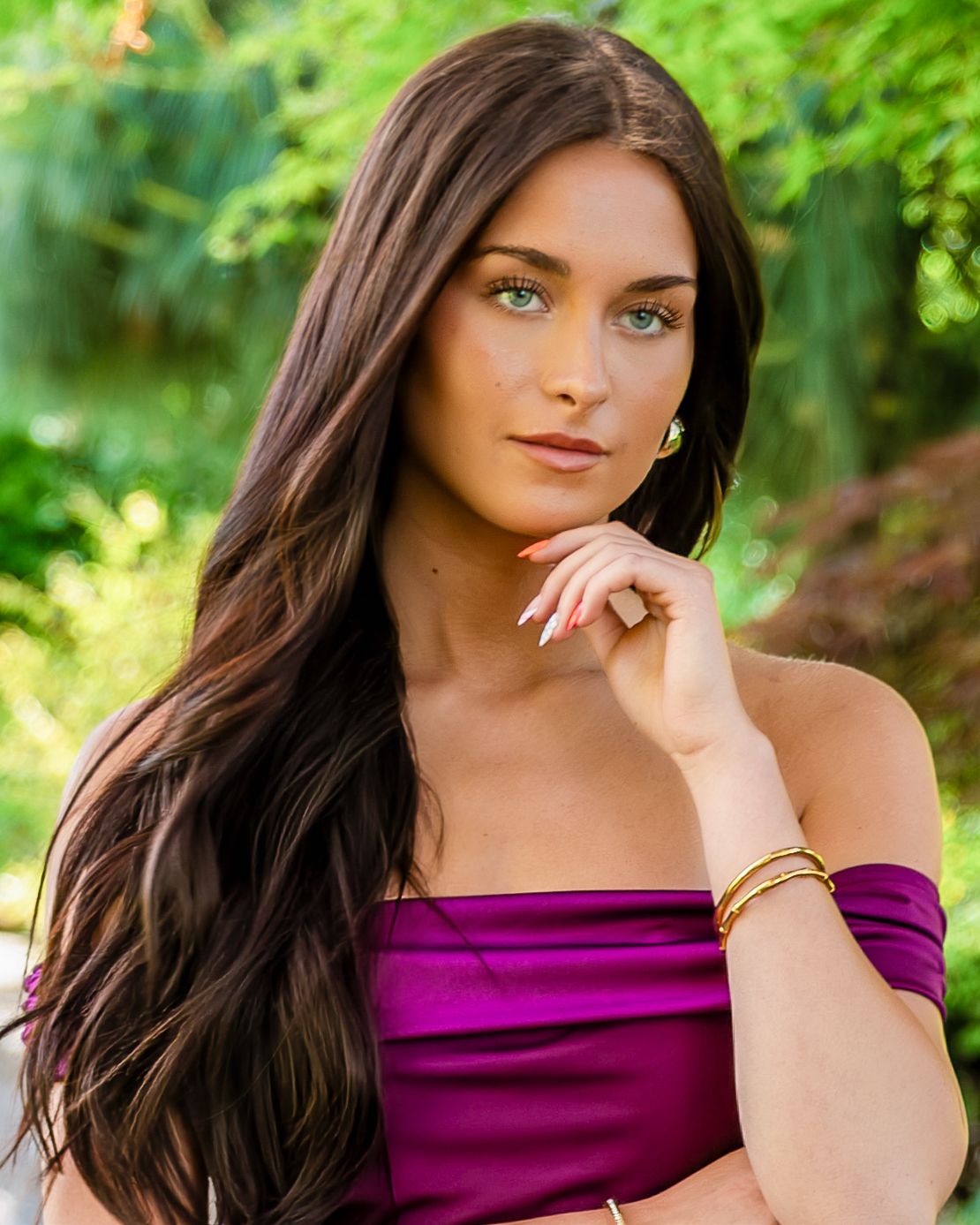 Young woman with green eyes long dark hair wearing purple off-shoulder top outdoors. Senior portrait