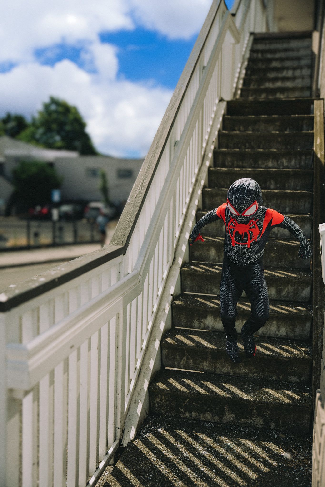Person in Spider-Man costume climbing outdoor stairs with blue sky and clouds in background.