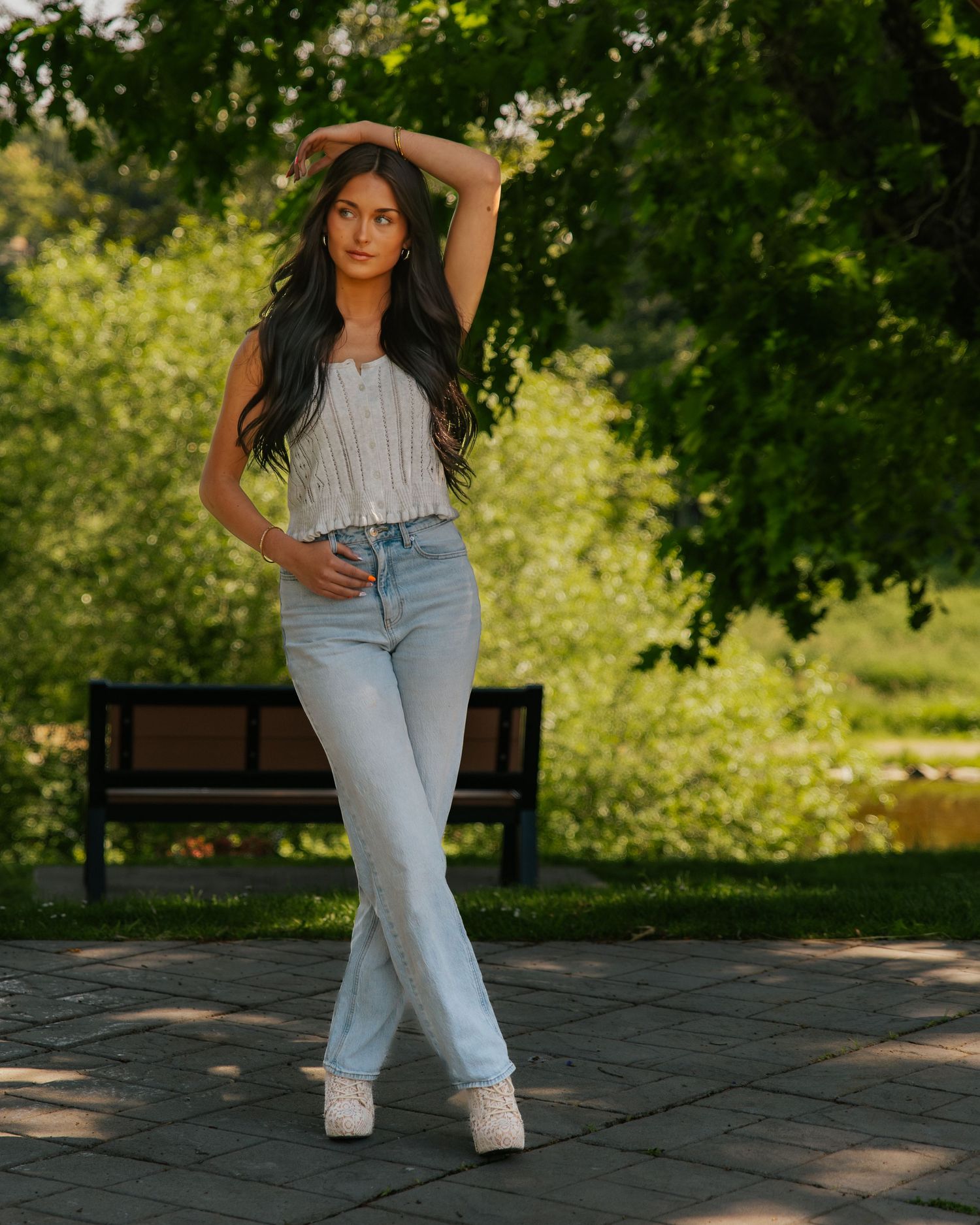 Young woman in light jeans and white top posing gracefully in a sunny green park with a bench in the background.