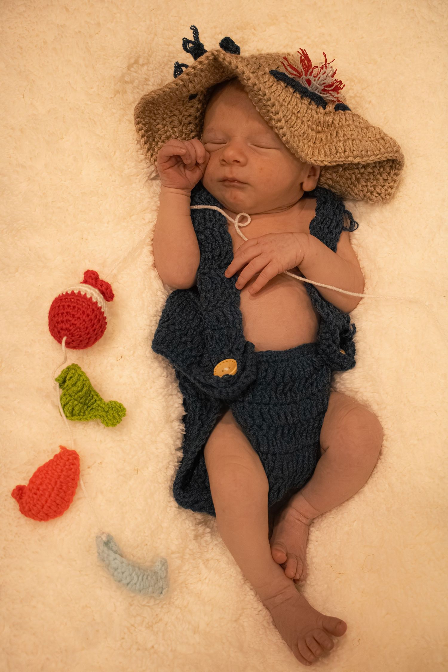 Sleeping newborn baby dressed in crocheted fishing overalls and hat, surrounded by colorful crocheted fish toys.