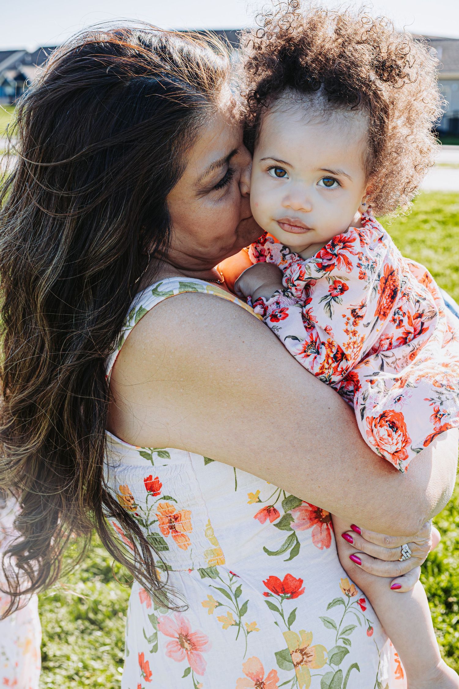 A mother in a floral dress kisses her curly-haired toddler outdoors on a sunny day.