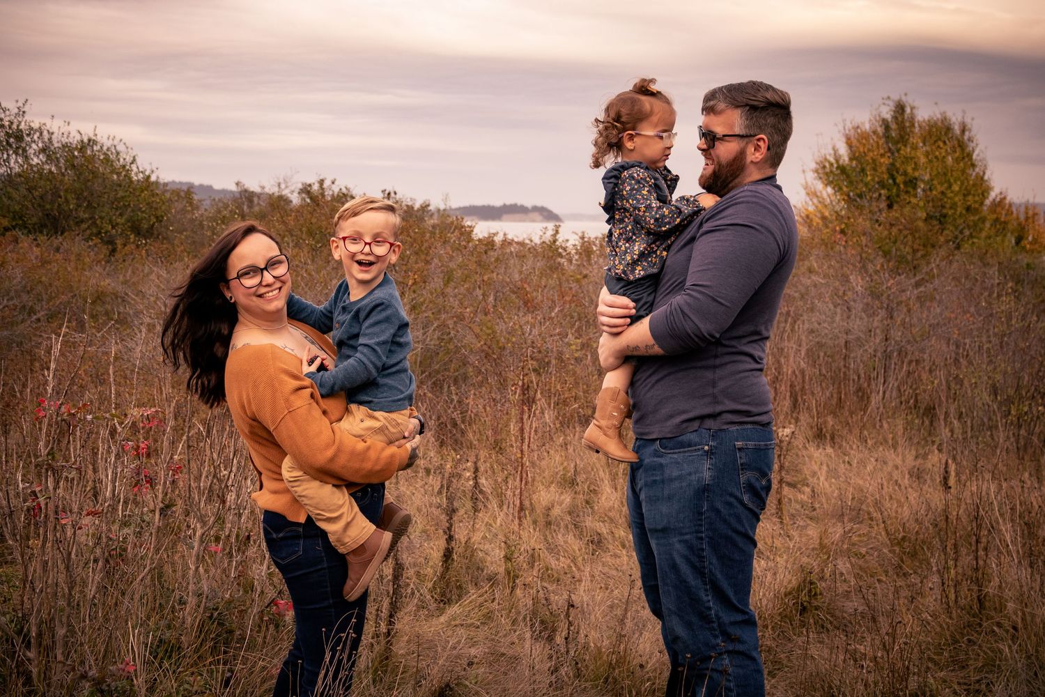 Happy family of four playing in a fall meadow with parents lifting laughing children outdoors.