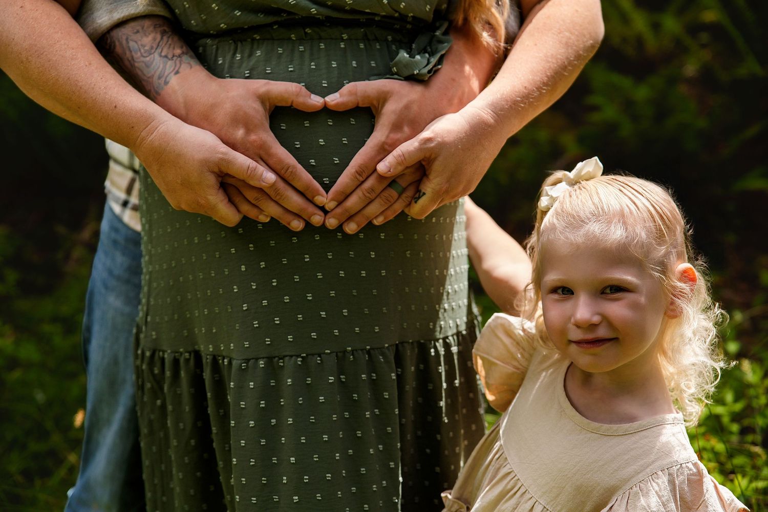 Pregnant woman in green dress with partner making heart shape on baby bump, toddler girl smiling beside them outdoors.