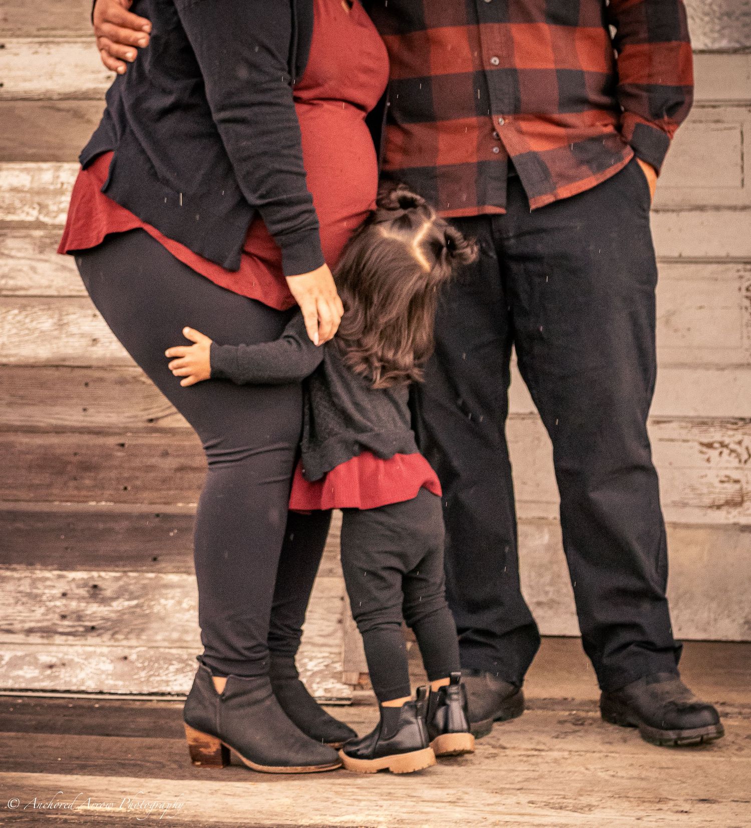 Pregnant family maternity photo with toddler hugging mom's belly, wearing matching red and black outfits on rustic wood backdrop.