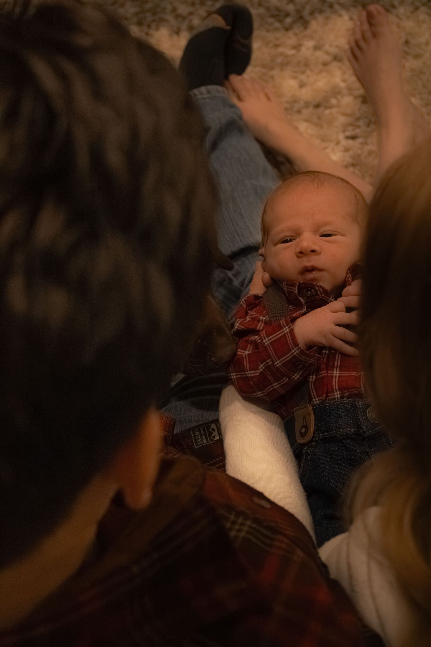 Newborn baby in red plaid shirt and jeans lying on carpet surrounded by family members looking down lovingly.