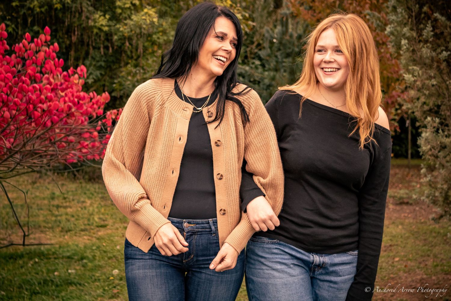 Two women smiling and laughing together outdoors in a garden with colorful autumn foliage in the background.