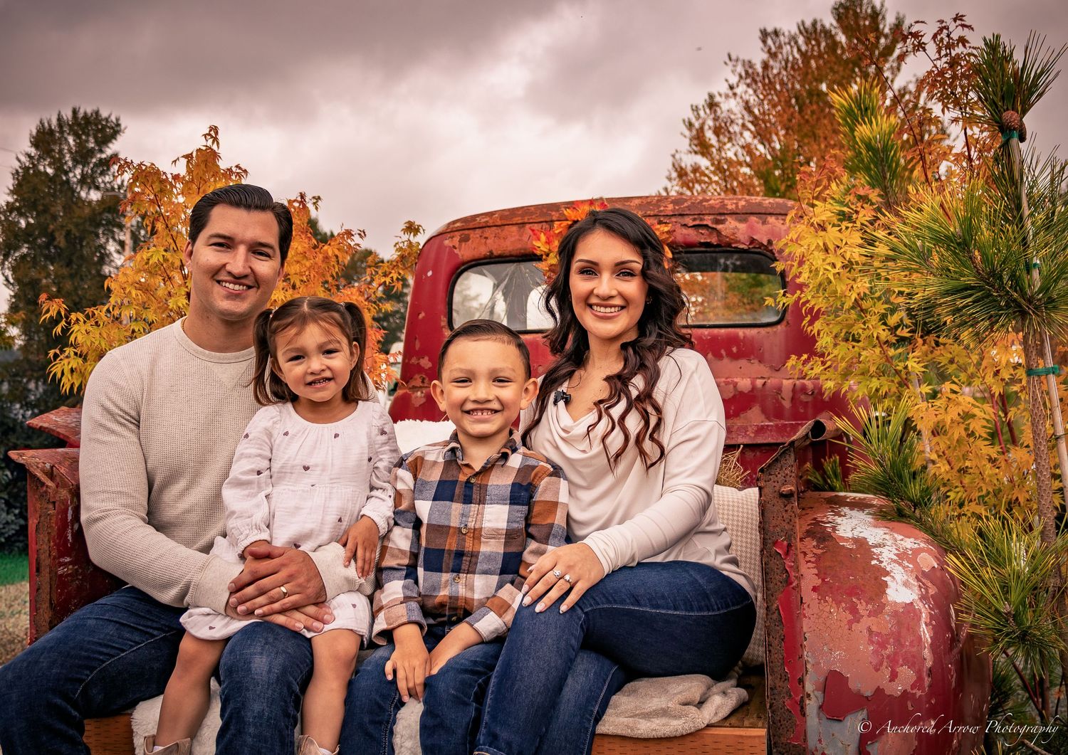 A happy family of four poses together on a vintage red truck surrounded by fall foliage.