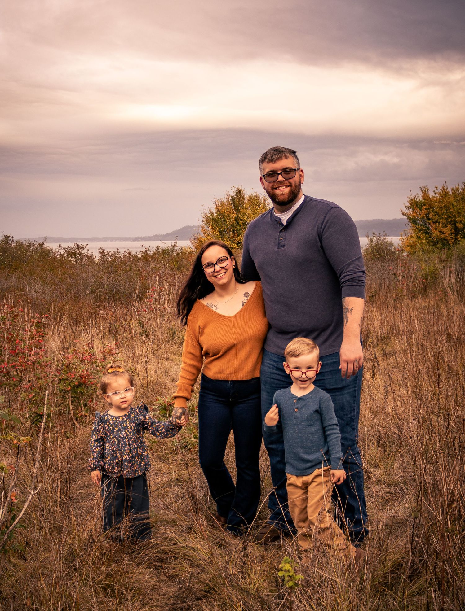 Family of four posing in golden autumn field with dramatic cloudy sky and water in the background.