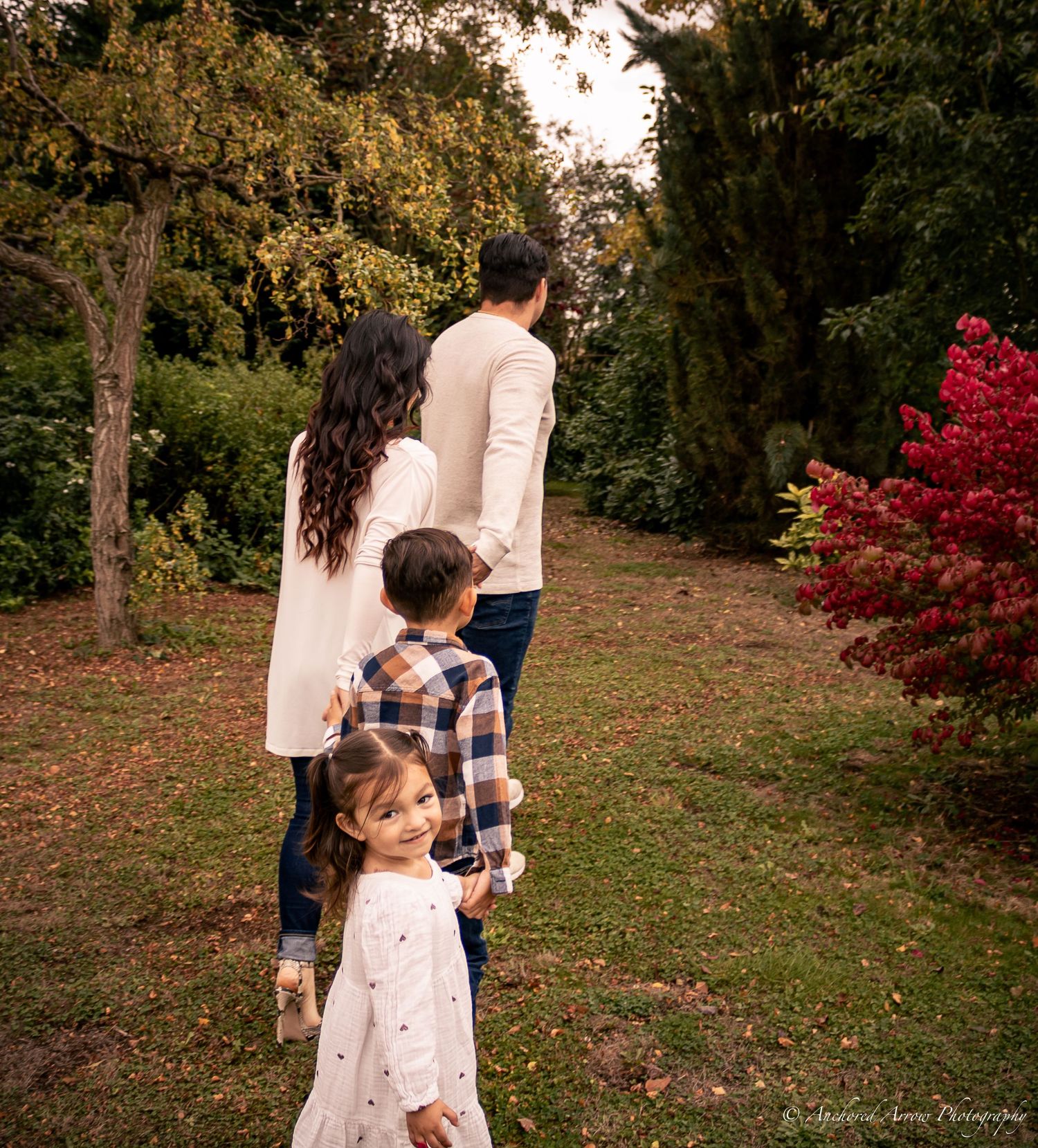 A family of four walks together through a lush garden with colorful autumn foliage, viewed from behind.