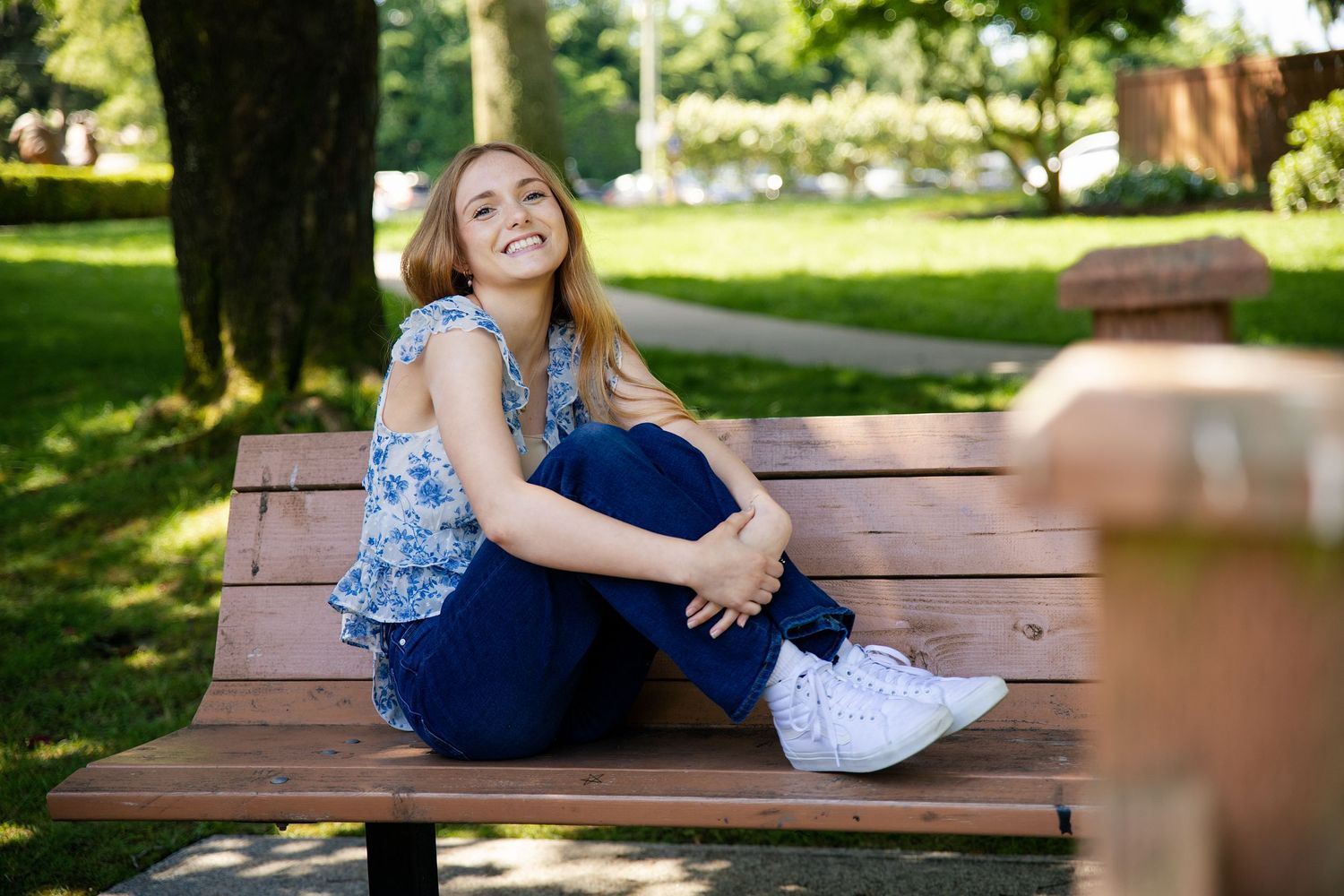 Young blonde woman smiling on a wooden park bench, wearing blue jeans and floral top on a sunny day.