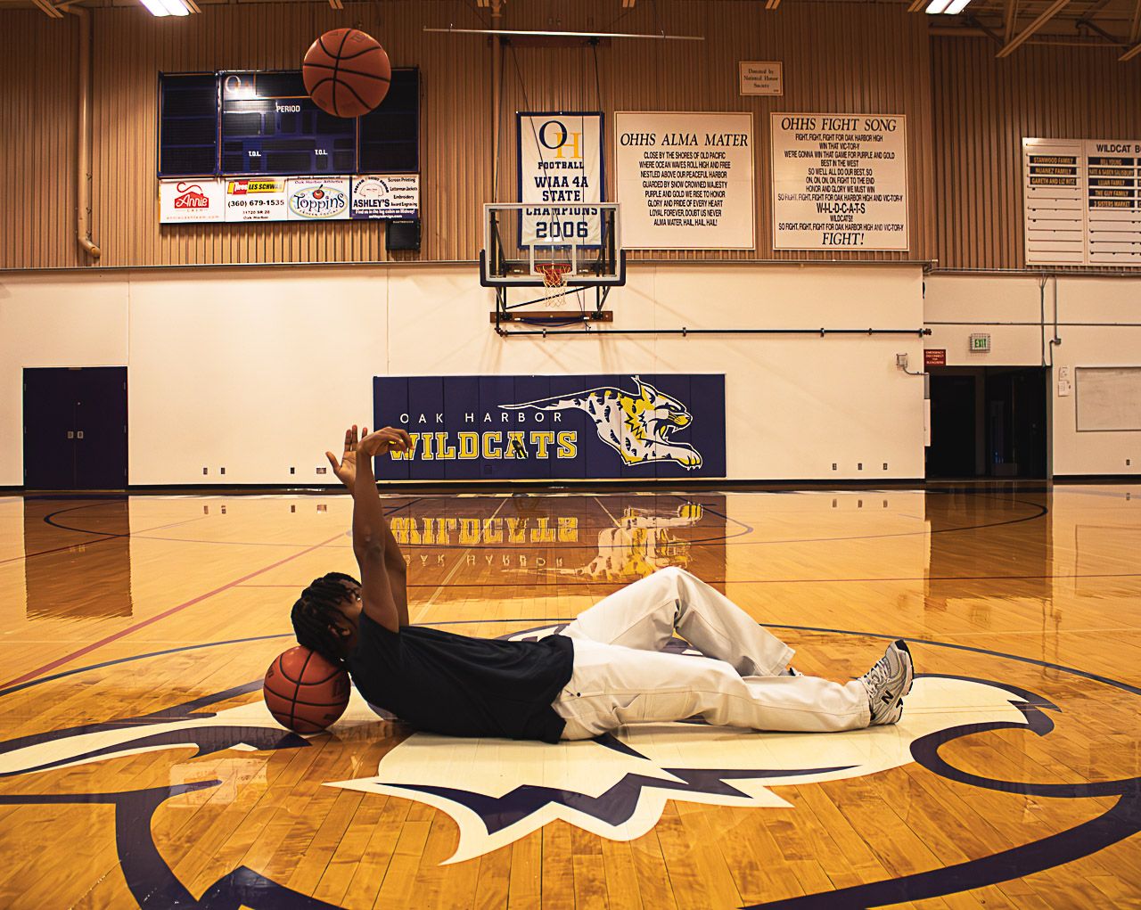 A person lies on a basketball court floor tossing a ball up in a school gymnasium.