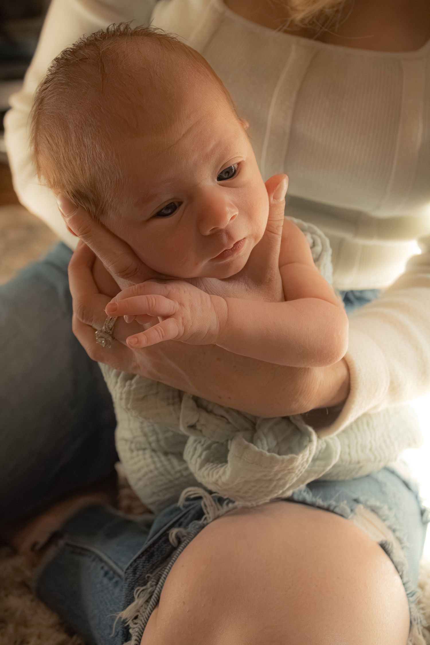 Alert newborn baby with blue eyes held gently in mothers arms in warm light.