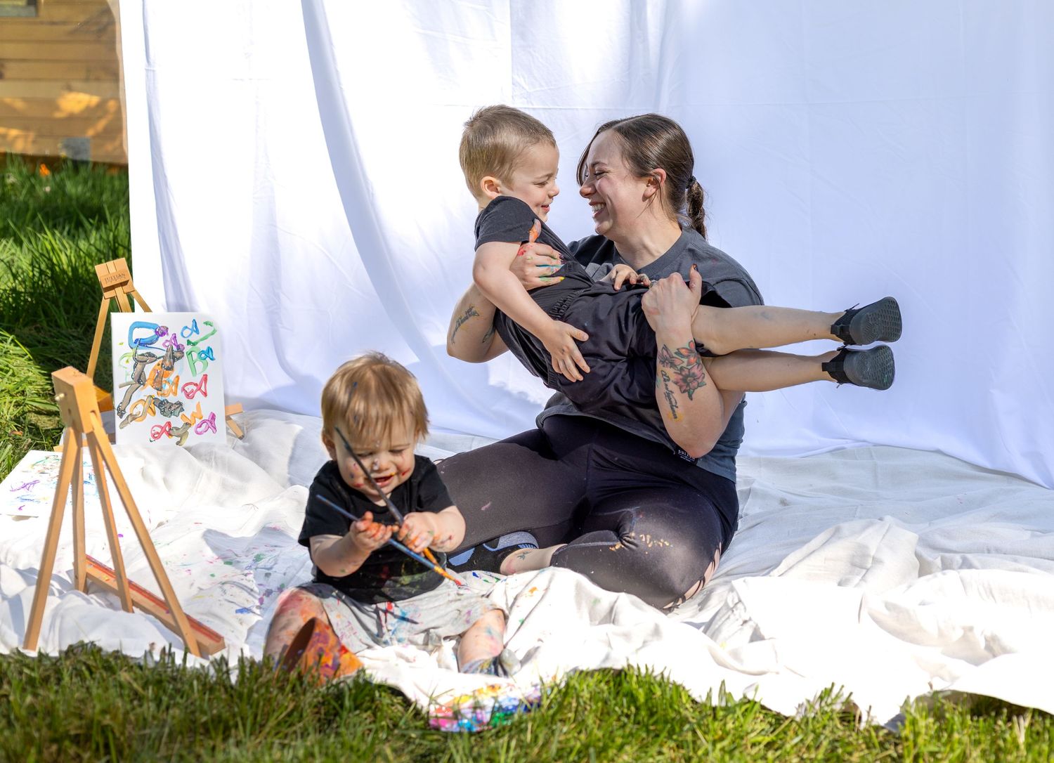 A mother sits on a white blanket outdoors, holding a baby while a toddler plays with paint beside an easel.