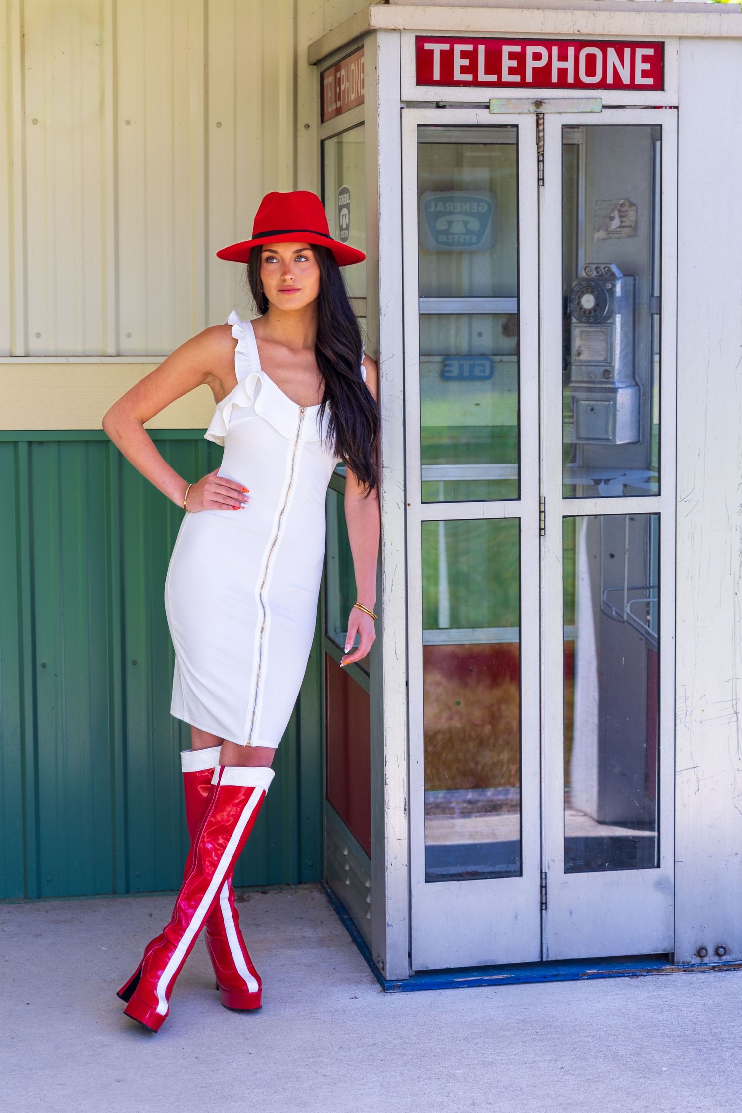 Woman in white dress, red hat and boots posing by vintage telephone booth outdoors.