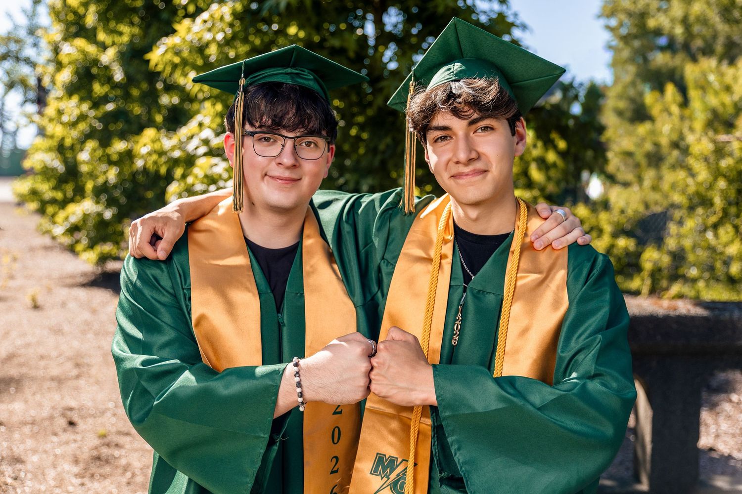 Two graduates in green and gold caps and gowns smile together outdoors at a graduation ceremony.