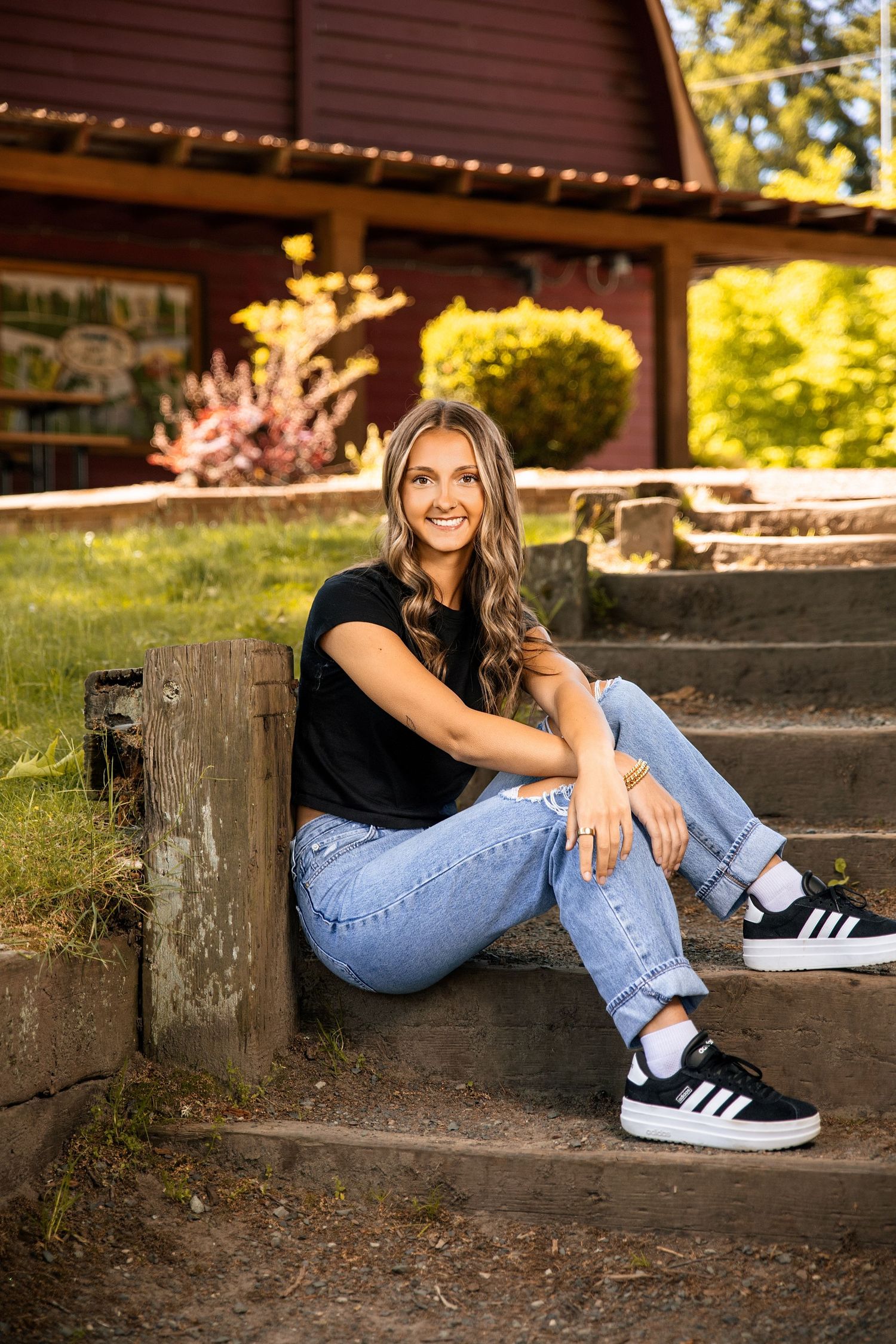Young woman smiling in black tee, jeans, and Adidas sneakers, sitting on stone steps outside a red barn.