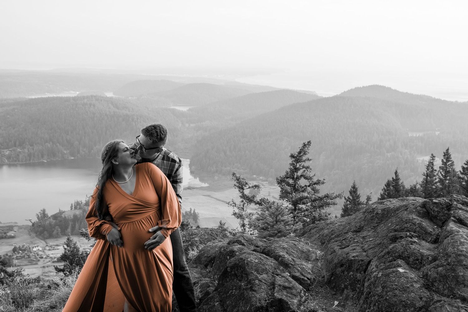A couple in orange dress viewing a scenic mountain landscape with pine trees and lake in black and white background.
