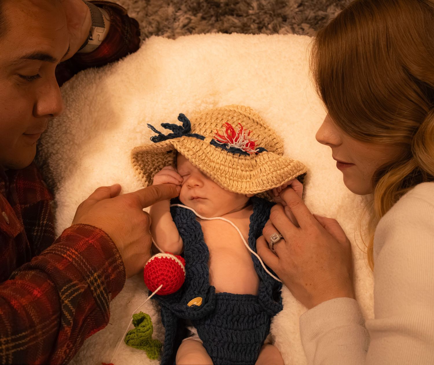 Newborn baby in crochet sun hat and denim overalls outfit, cradled by parents on white blanket.