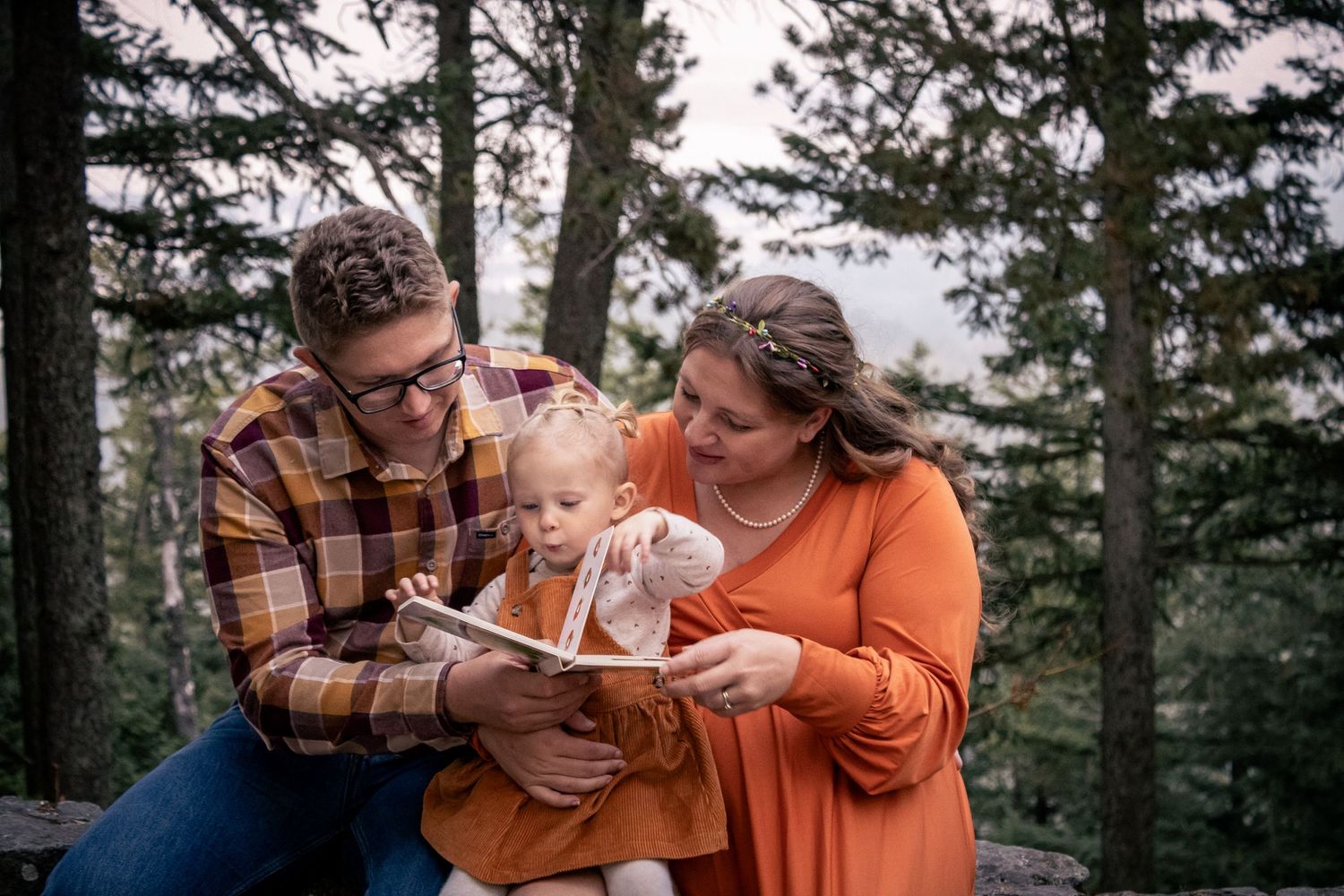 Family of three reading a book together outdoors among pine trees during a fall photo session.