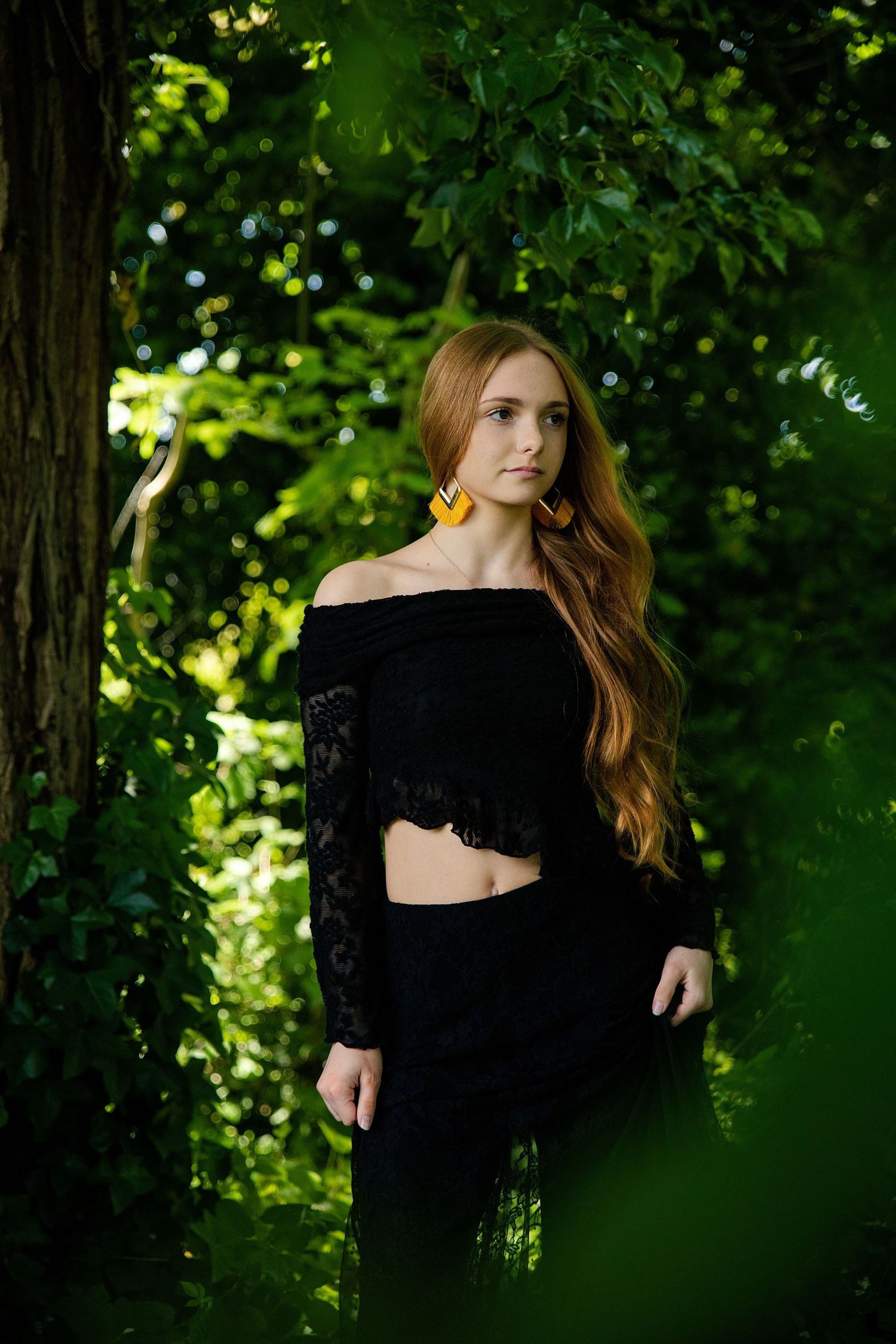 Young woman with long red hair in black lace off-shoulder crop top and skirt, wearing yellow earrings, posing outdoors.