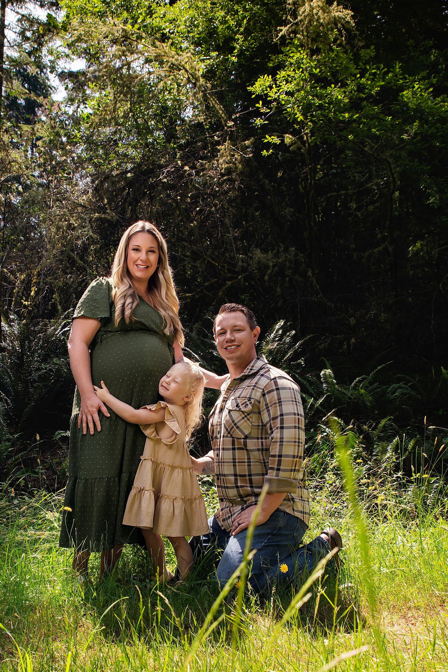 Pregnant woman in green dress with partner and young daughter posing in sunlit forest clearing for maternity photo.