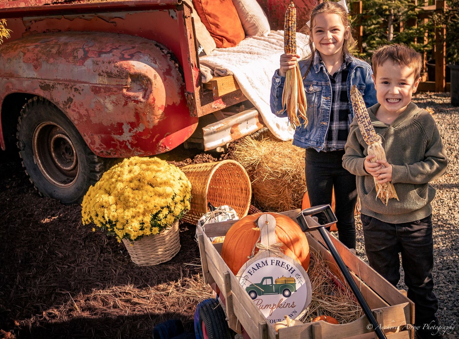 Two children pose with fall harvest decorations, pumpkins, hay bales, and mums beside a vintage red truck. Fall Mini.