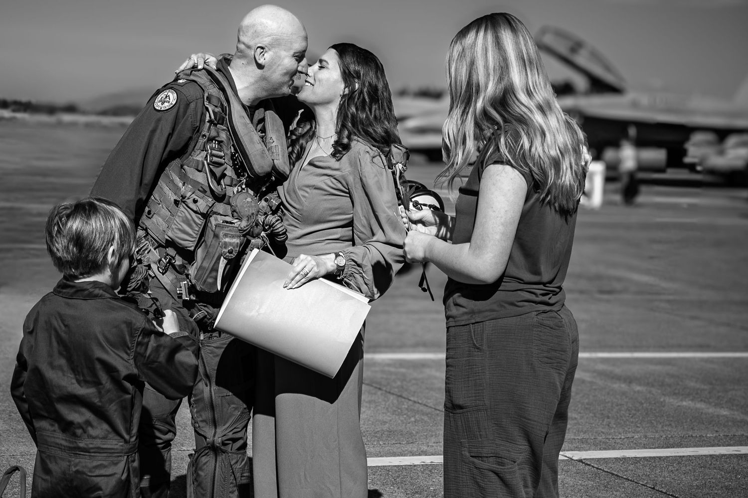 Military pilot in flight suit kissing spouse on airport tarmac during homecoming reunion.