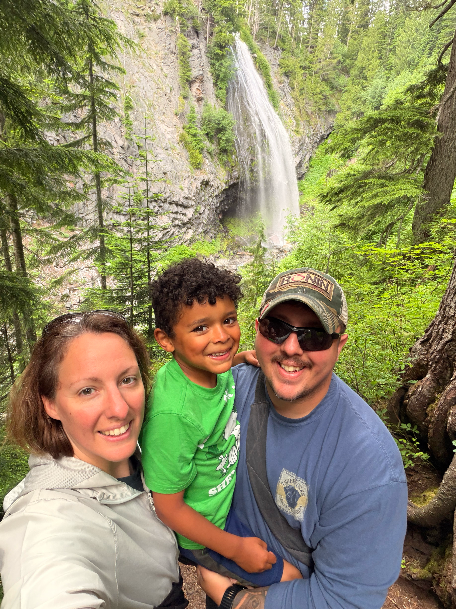 Family of three smiling together in front of a tall waterfall surrounded by lush green forest trees.
