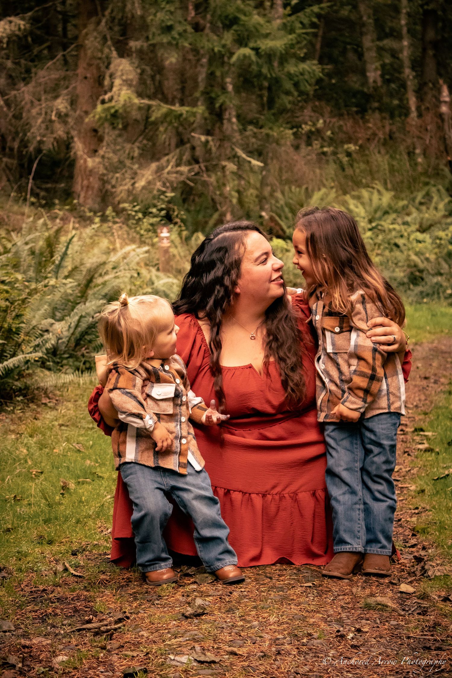 A mother in a red dress kneels outdoors smiling at her two young daughters in a forest setting.