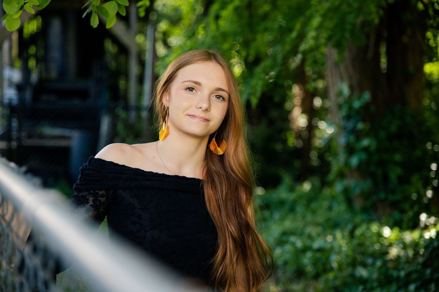 Young woman with long auburn hair wearing yellow earrings and black off-shoulder top poses outdoors.