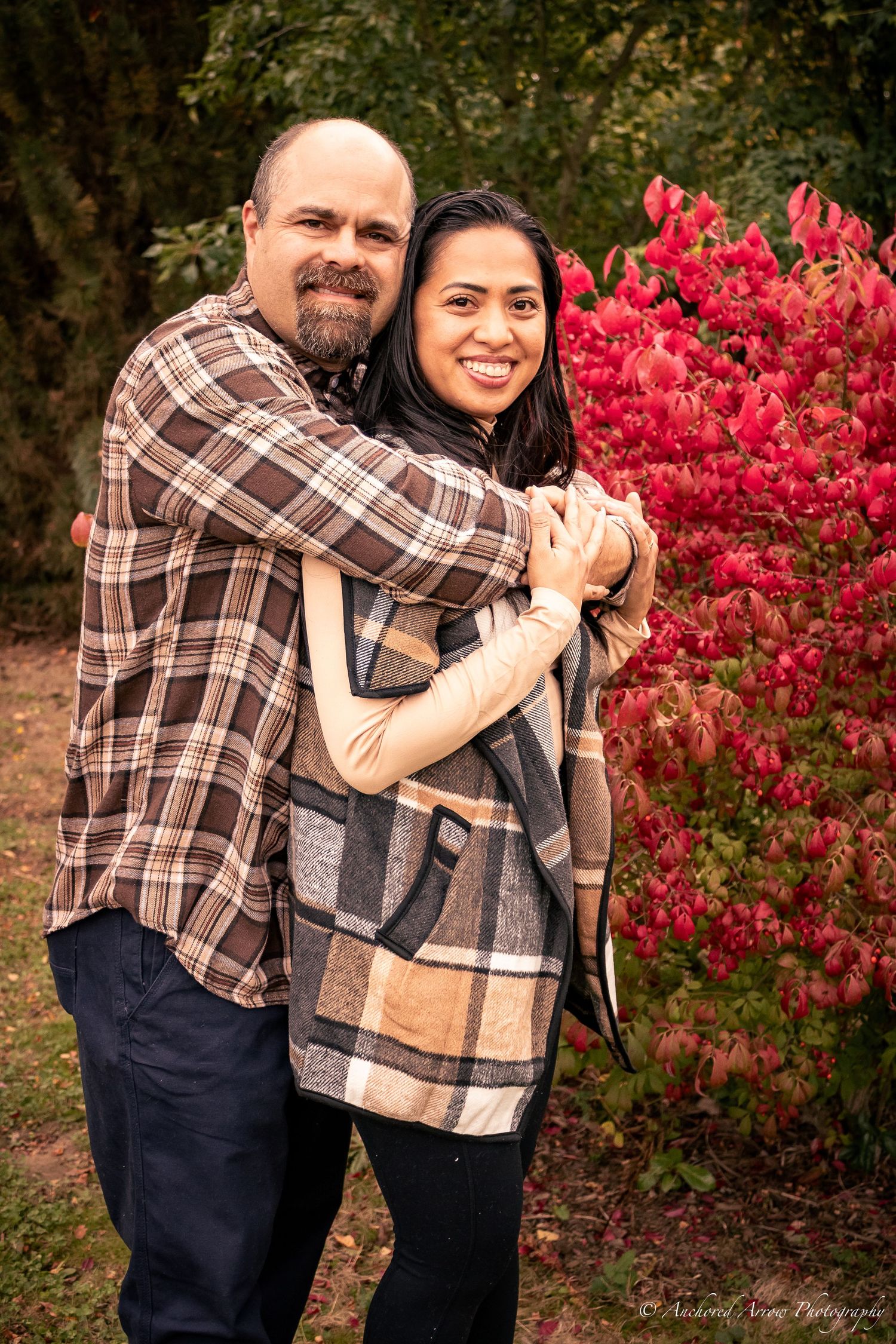 A smiling couple embraces warmly in front of vibrant red autumn shrubs in an outdoor setting.