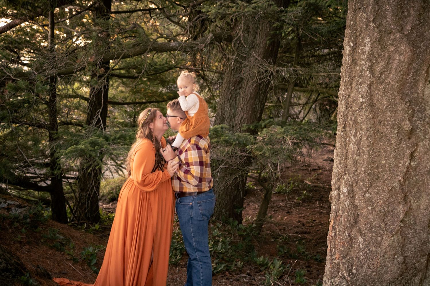 Family of three in autumn forest, mother in orange dress, father lifting baby among pine trees.