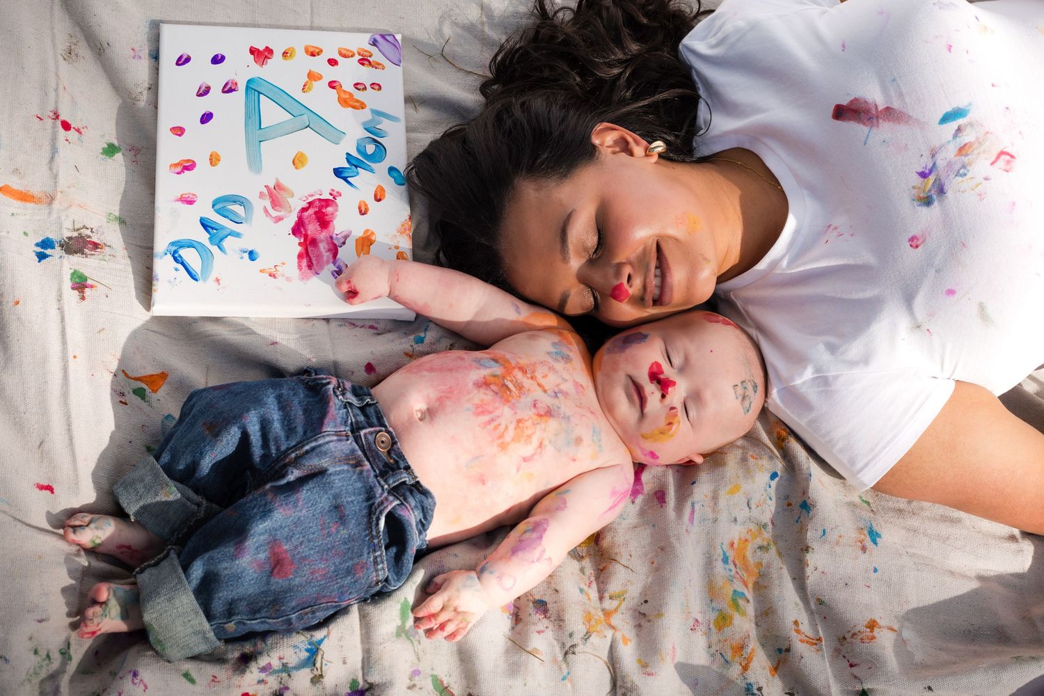 Mother and baby lying together covered in colorful paint marks, with a handmade card reading Dad and Mom nearby.