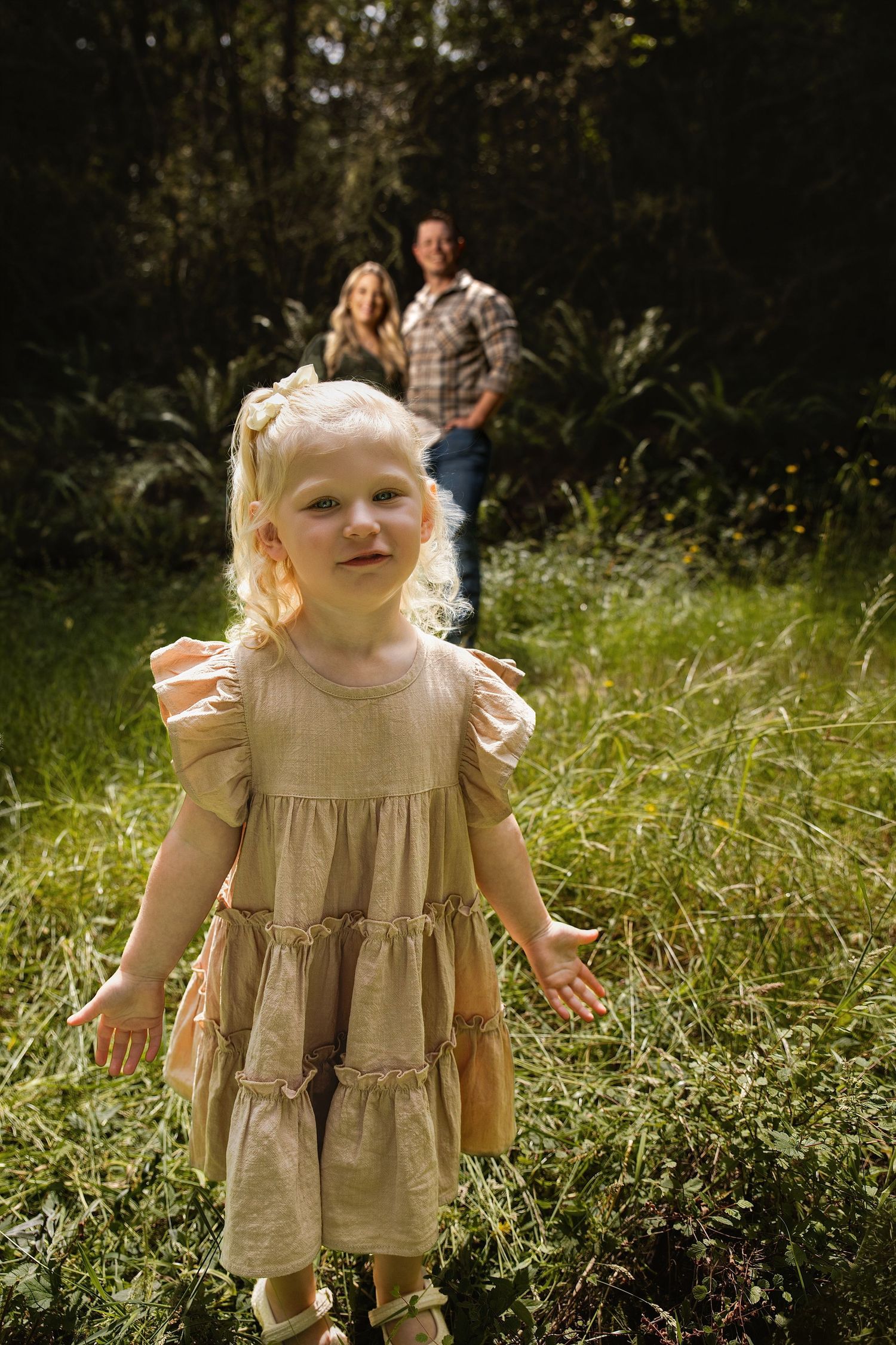 Young blonde girl in beige dress runs through green field with parents smiling in background.