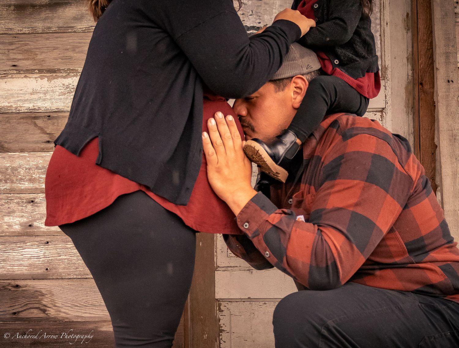 Person kneeling and kissing a pregnant woman's belly near a rustic wooden wall, captured by Sandoval Arena Photography.
