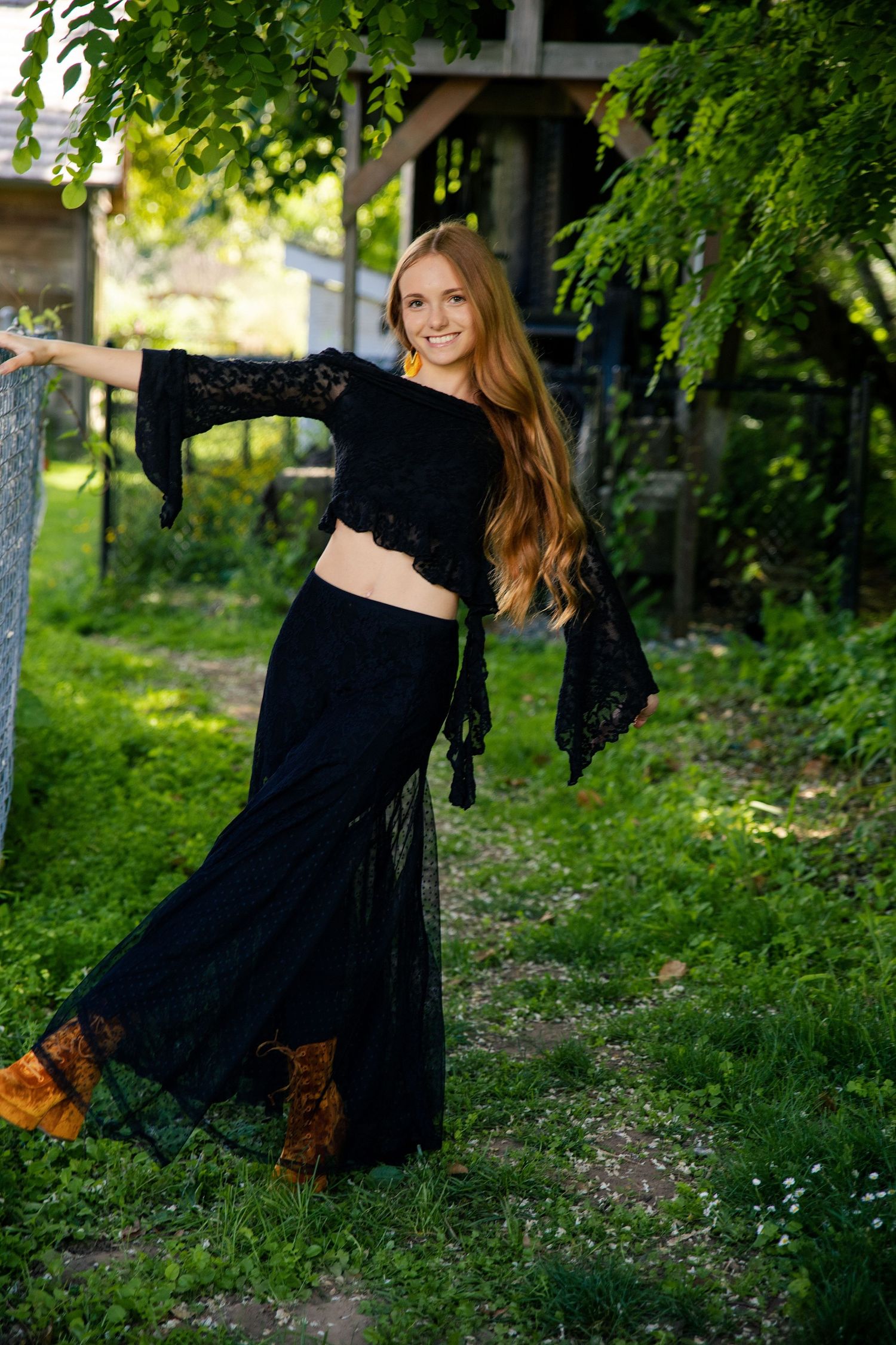 Smiling woman with long red hair wearing a black lace crop top and maxi skirt posing outdoors in a lush green garden.