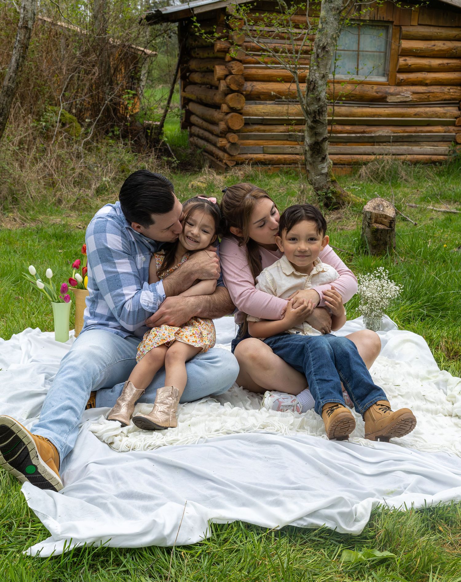 Happy family of four cuddling on a white blanket outdoors near a rustic log cabin in a lush green yard.
