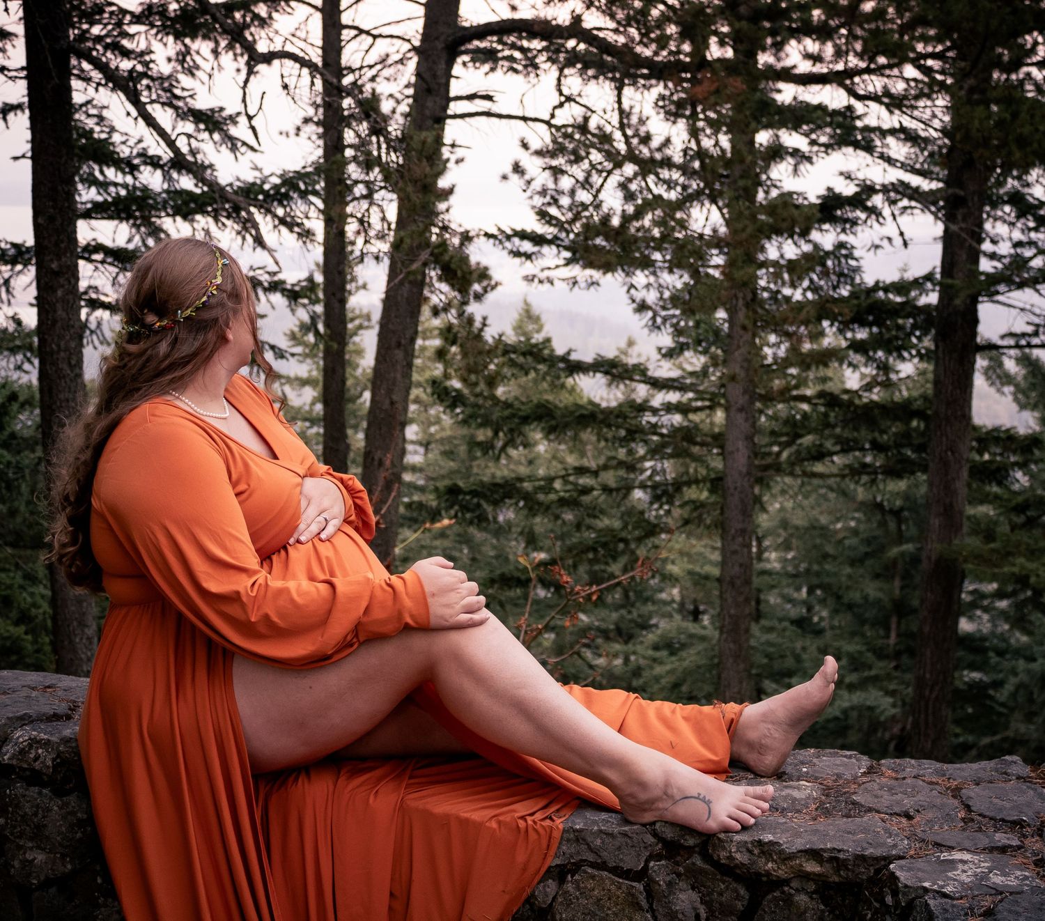 Pregnant woman in orange dress sitting on stone wall surrounded by trees.