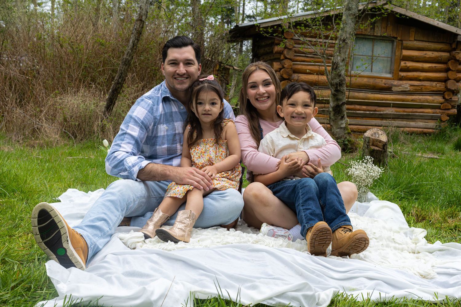 Happy family of four smiling together on a white blanket outdoors near a rustic log cabin in nature.