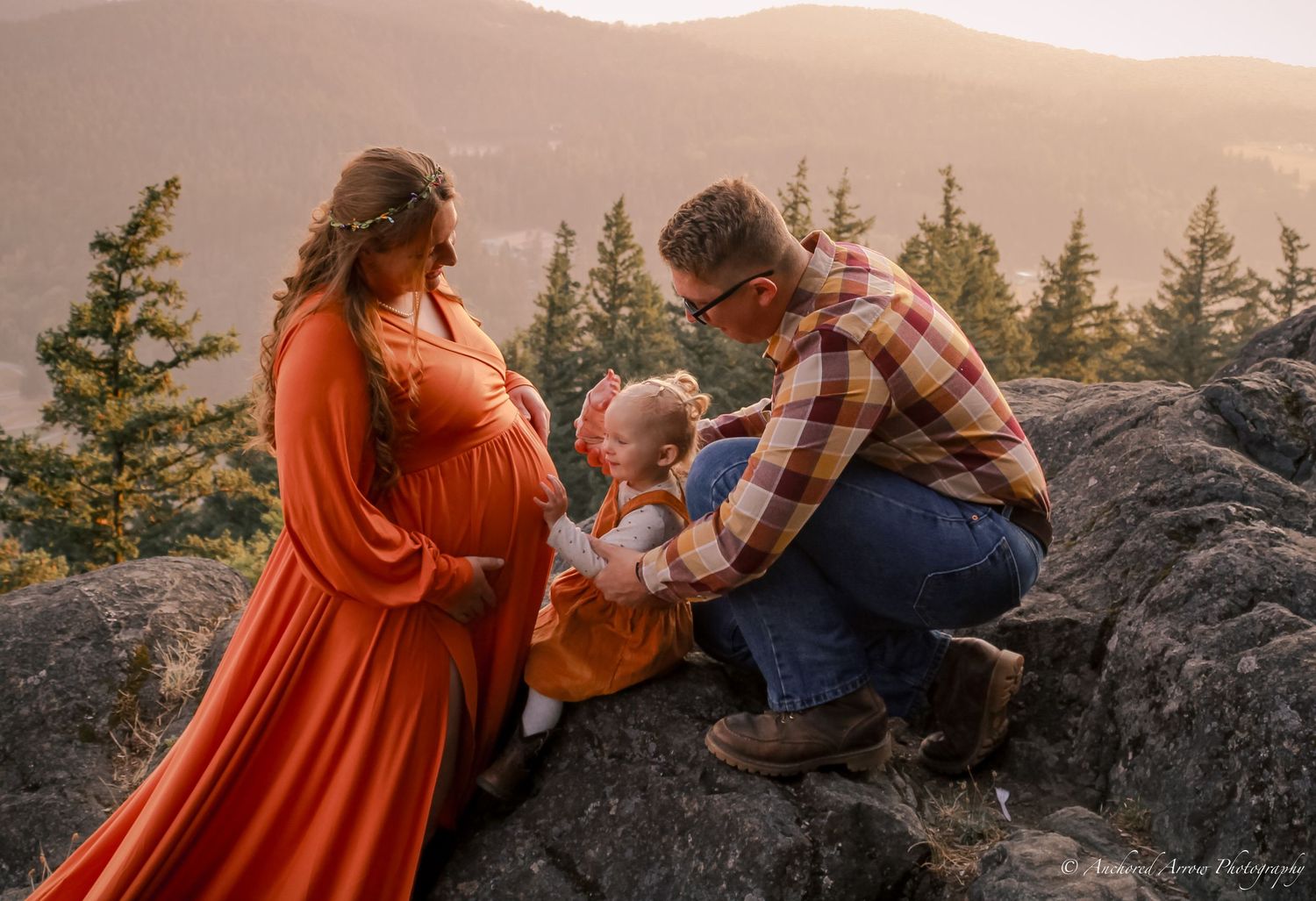 Pregnant woman in orange gown with partner and toddler share a tender moment on rocky mountain overlook at sunset.