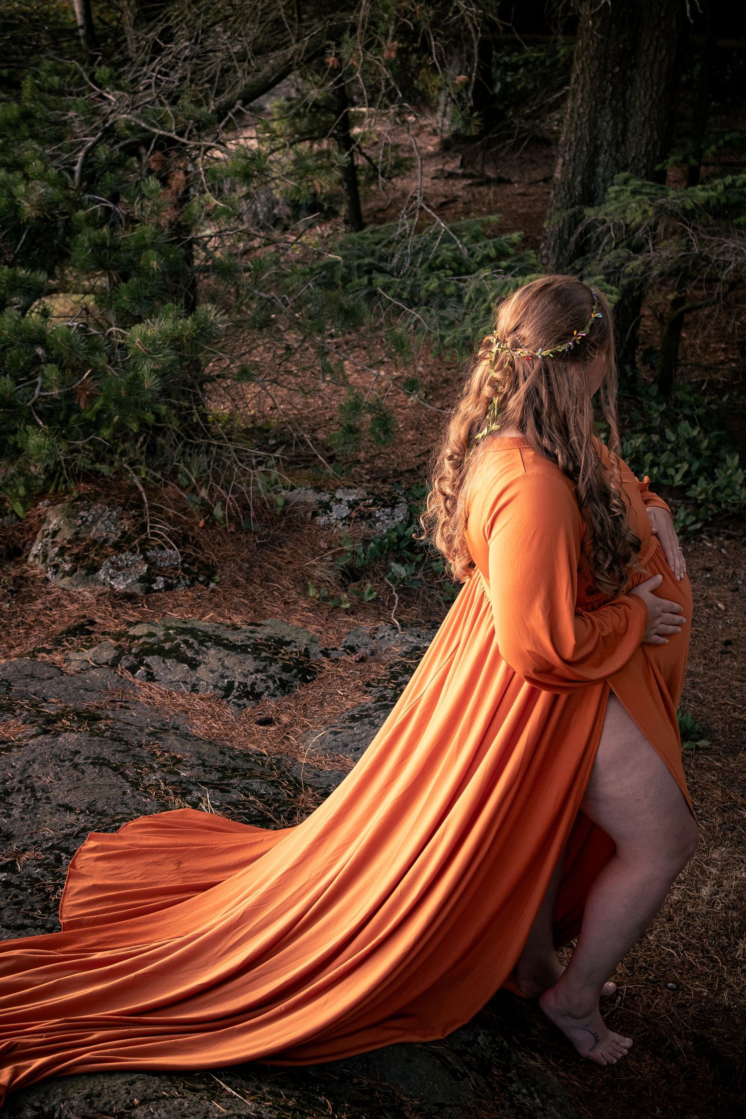 Pregnant woman in elegant orange flowing gown standing in a forest with a floral crown in her hair.