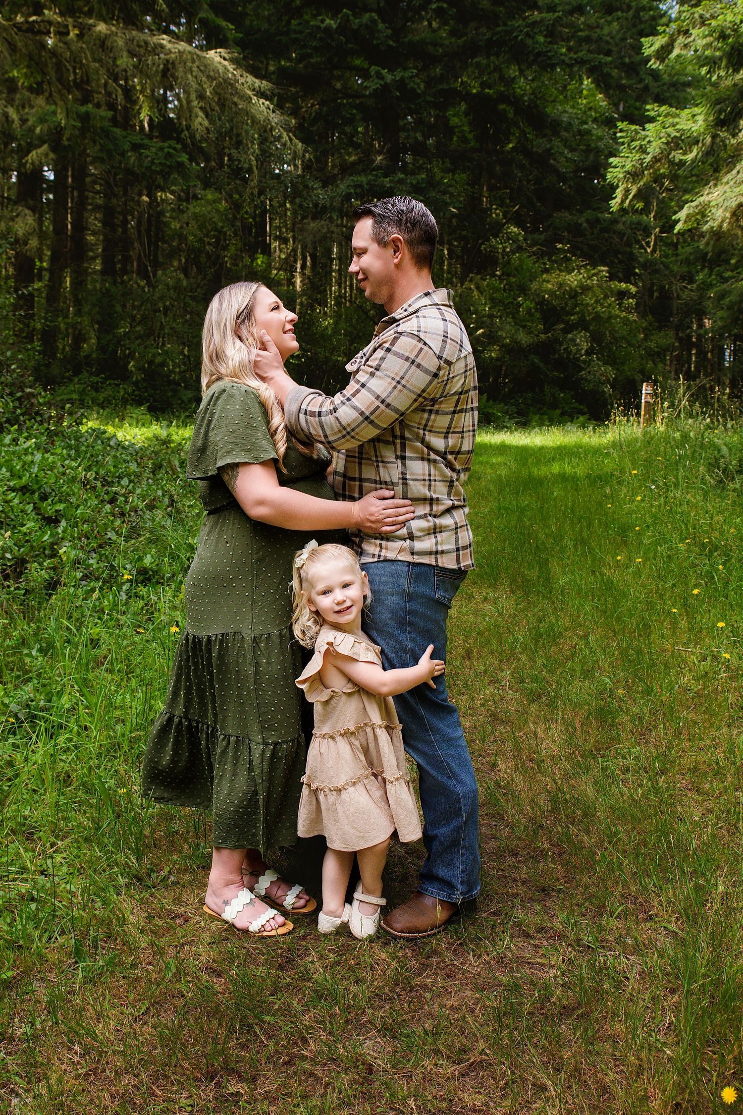 A happy family of three poses outdoors, parents gazing at each other while their toddler daughter stands between them.