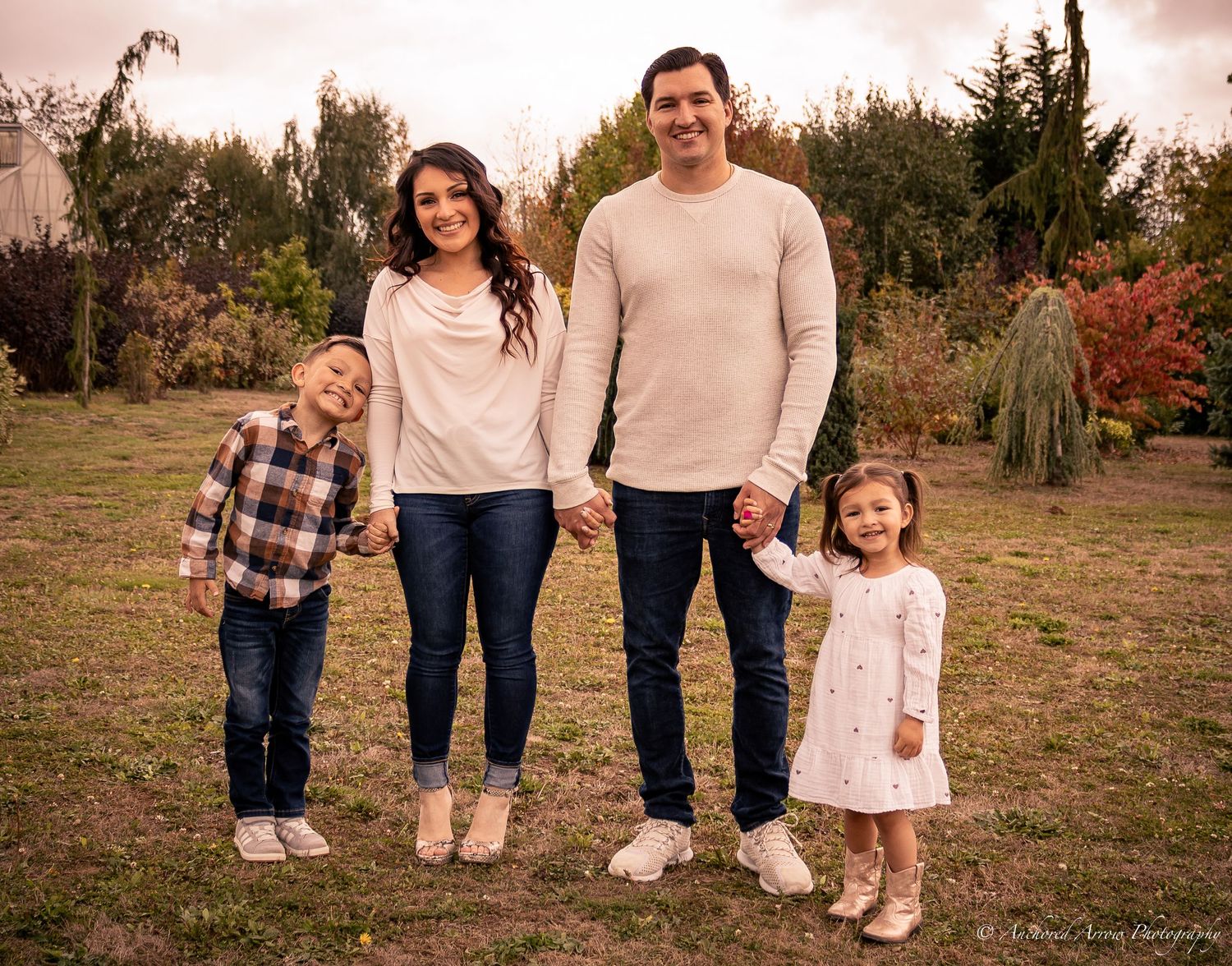 Family of four posing outdoors in a garden, parents holding hands with two young children in coordinated outfits.