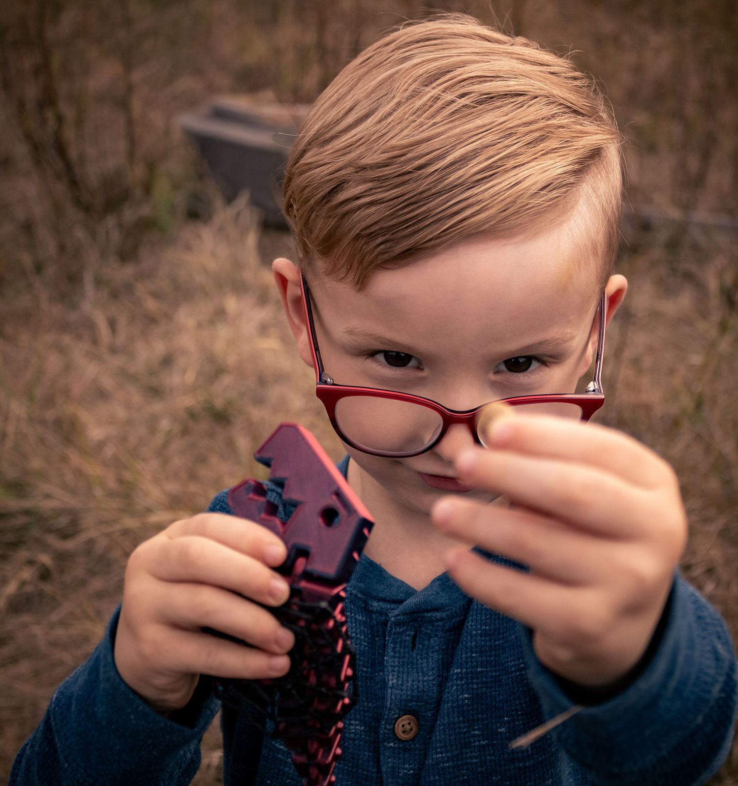 Young boy with red glasses examining a colorful toy outdoors among dry grass.