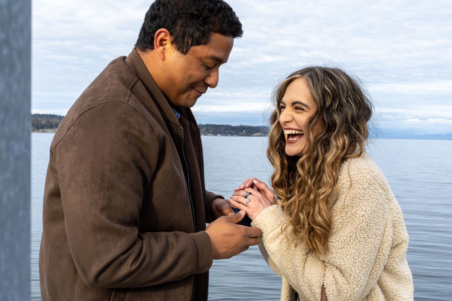 A man places a ring on a laughing woman's finger during a romantic waterfront proposal.